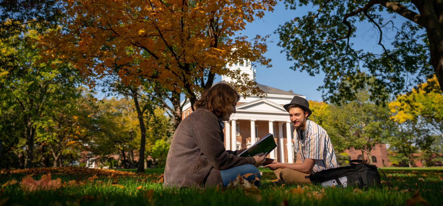 Two students study together outside under the bright colors of autumn trees.