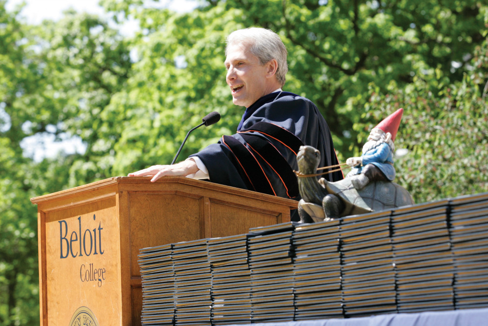 President Scott Bierman speaking at 2012 commencement flanked by a garden gnome riding a turtle.